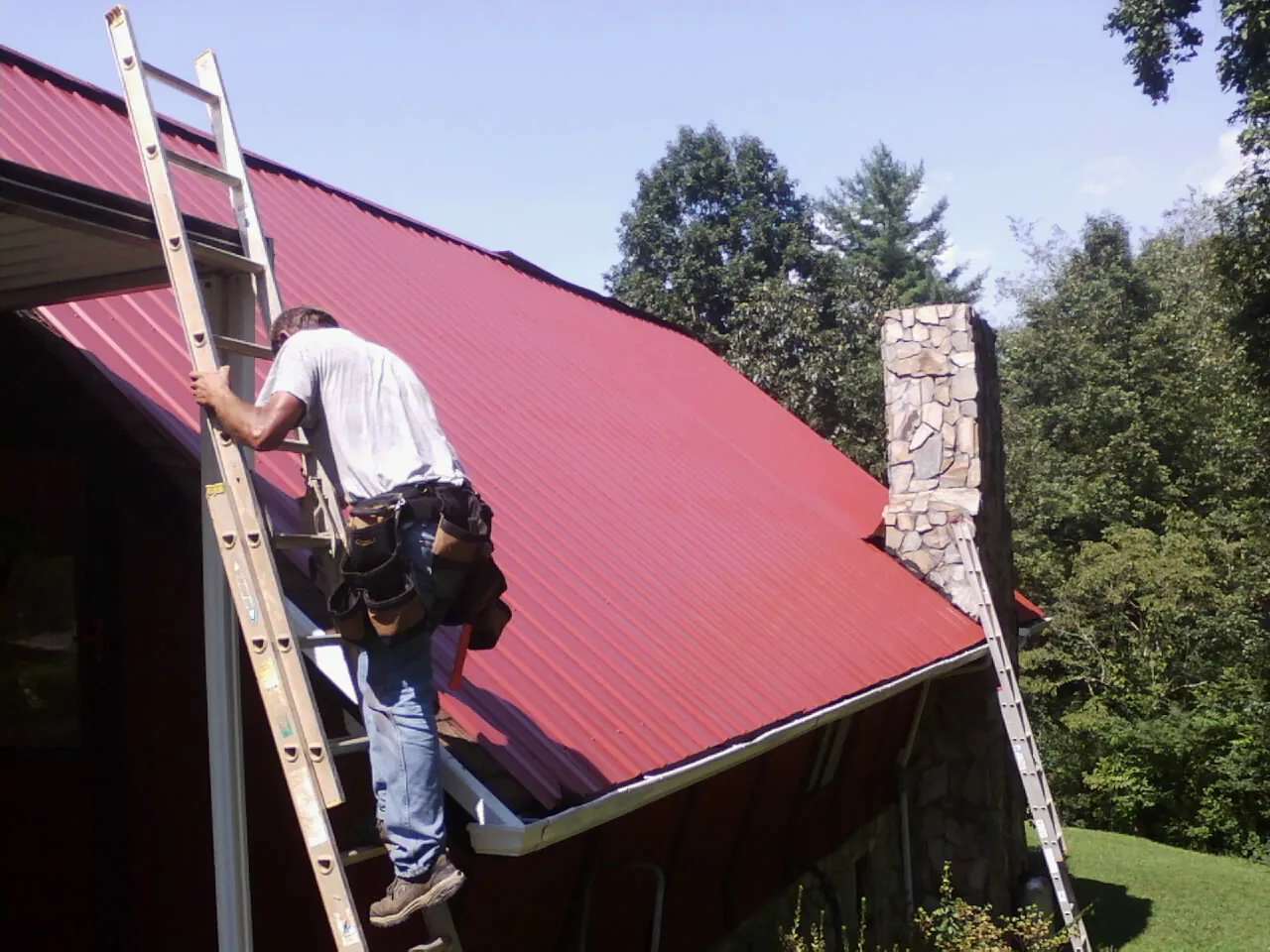 Roofer on a ladder beside a red metal roof and stone chimney — Soffit & Fascia Repair by Roofing Brothers in Leverett